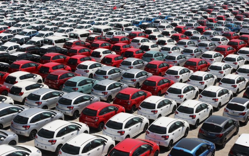 Vista aérea de carros novos de diferentes cores estacionados em fileiras em um grande pátio.